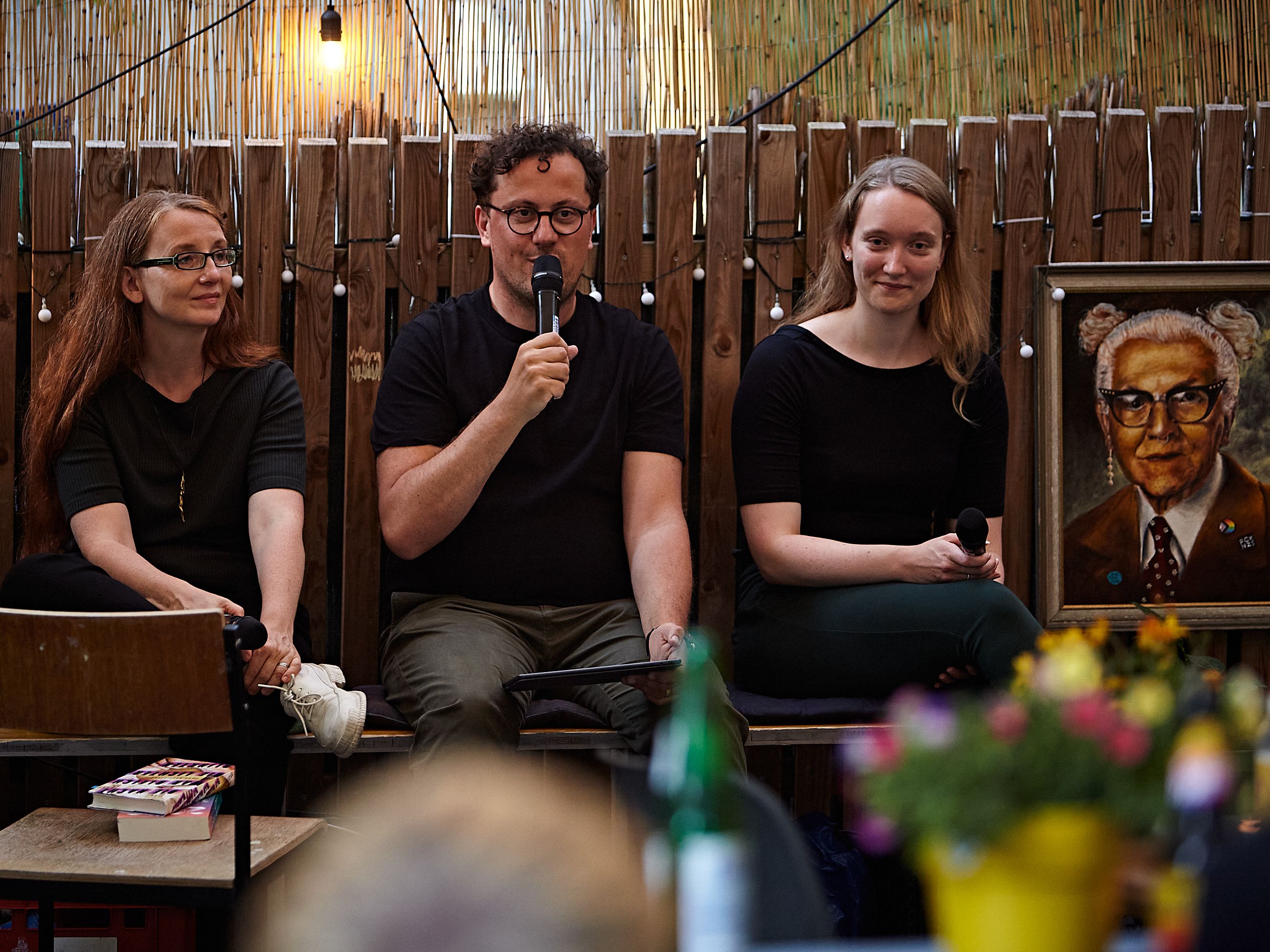 Drei Menschen sitzen vor einem Publikum, einer spricht in ein Mikrofon