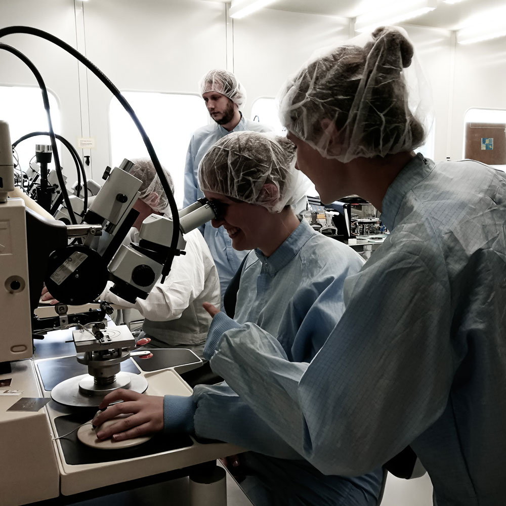 Studierende untersuchen in einem Reinraum Mikrosysteme unter einem Mikroskop.