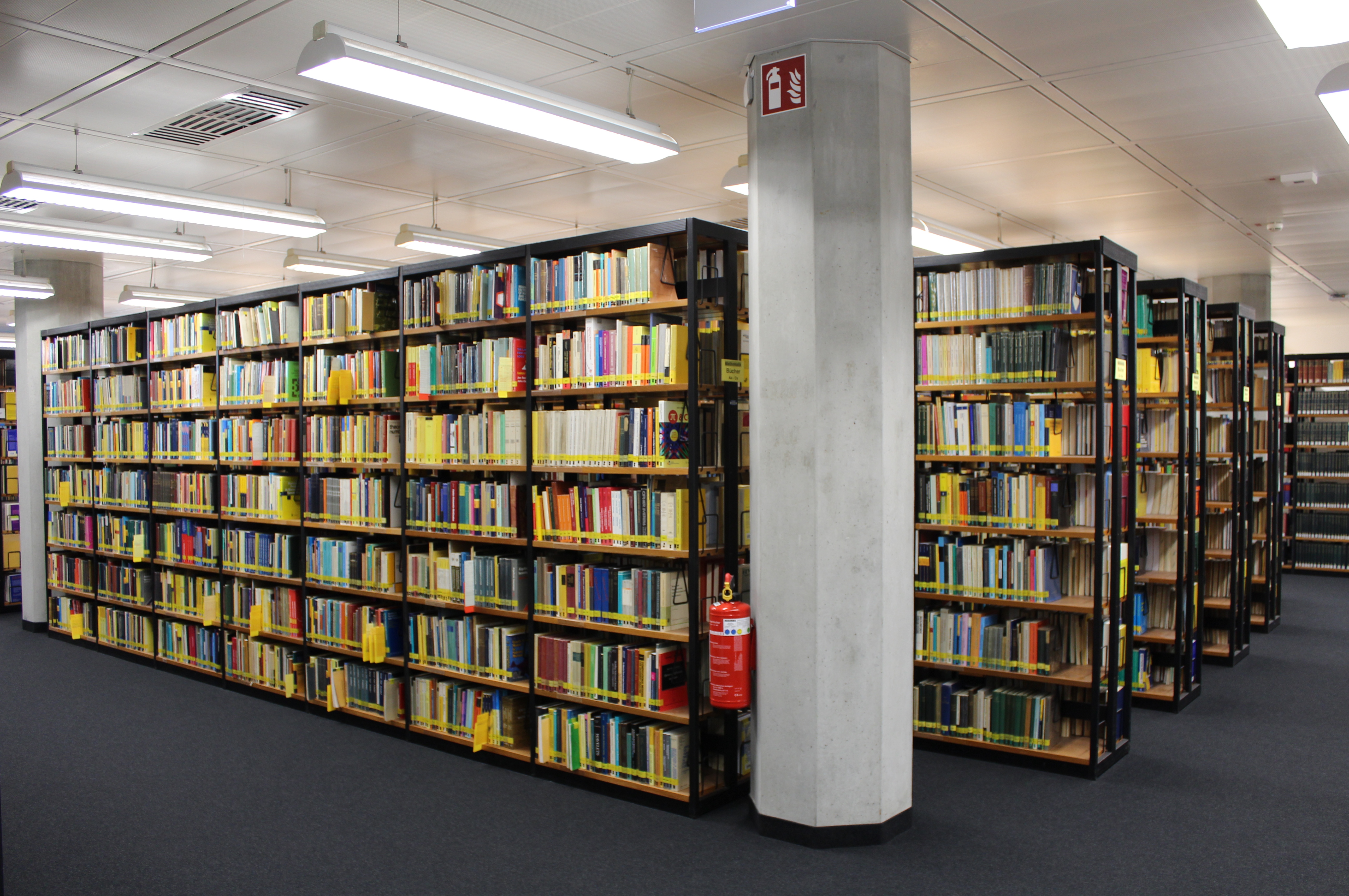 Several shelves are filled with books, a fire extinguisher can be seen on a column next to the shelf.
