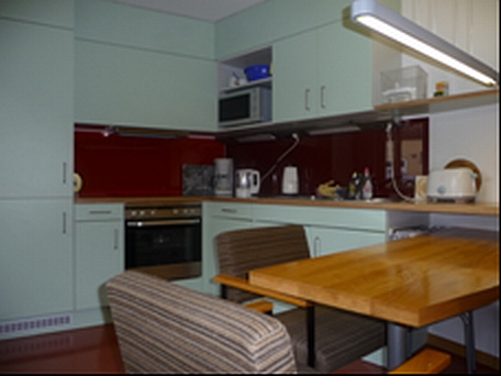Image: common kitchen with a table and chairs in the foreground, a kitchen unit mit a stove a fridge and several kitchen appliances