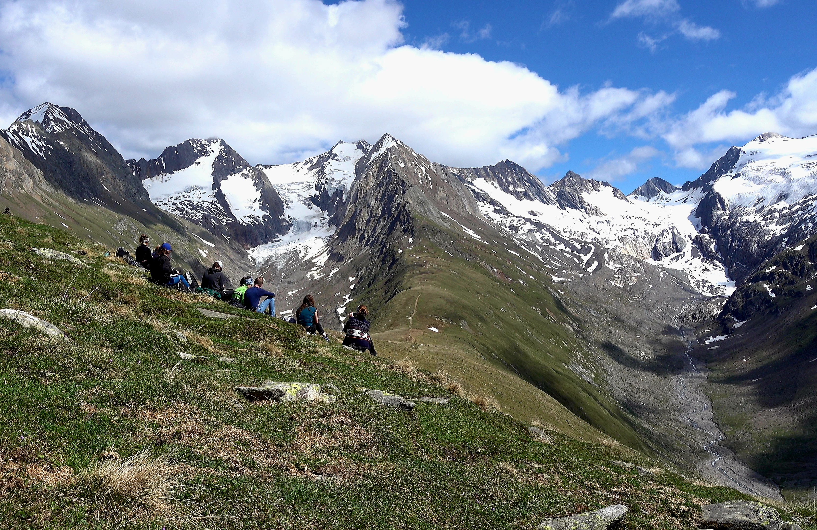 Auf dem Bild sieht man das Gebrige der Alpen und die Exkursionsteilnehmen, die dort auf der Wiese sitzen