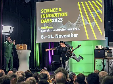 Blick auf Bühne über Publikum, auf der Bühne Mann im grünen Anzug und Mann mit Gitarre, der ein Bein nach hinten in die Luft streckt