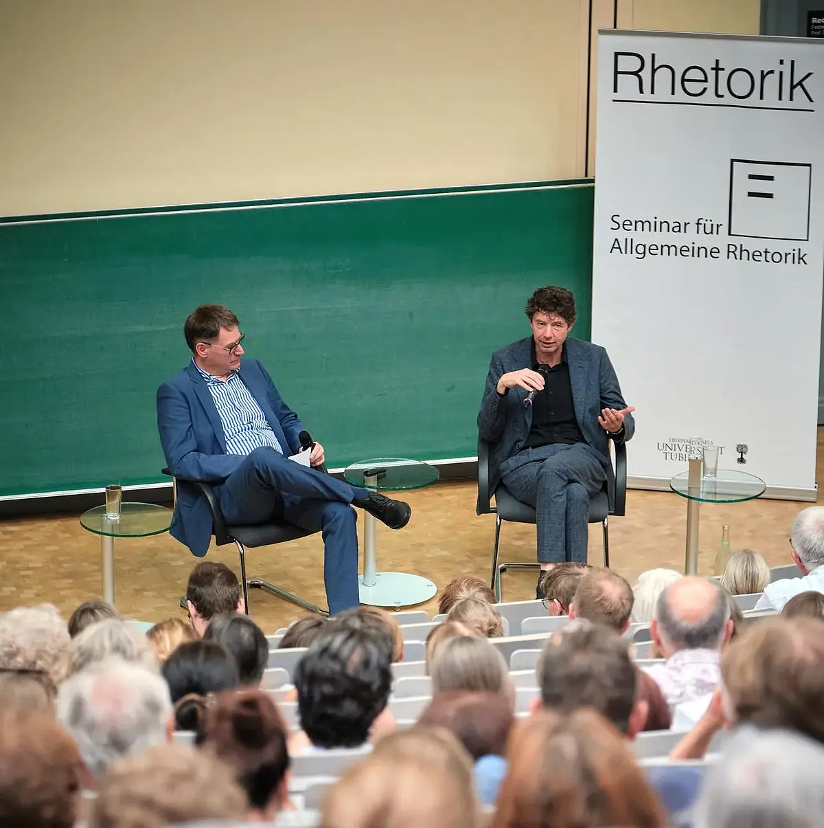 Olaf Kramer und Christian Drosten sitzend im Gespräch, aus dem Publikum eines vollen Hörsaals gesehen