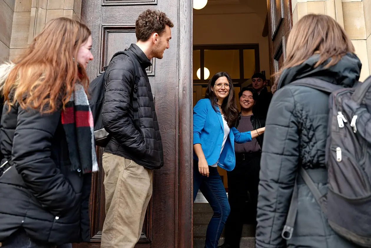 Foto von drei Personen draußen vor einer geöffneten Tür, die von einer Person aufgehalten wird. Eine weitere Person innen im Hintergrund.