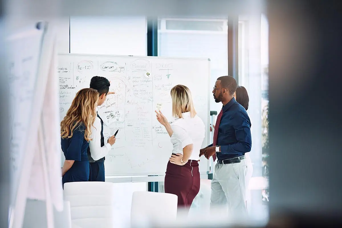 Vier Personen stehen mit dem Rücken zur Kamera vor einem beschriebenen Whiteboard und beraten darüber.