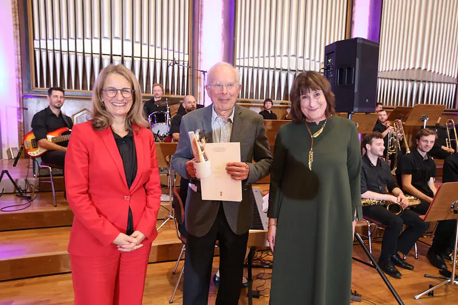 Professor Rainer Nagel hält seinen Preis in der Hand, links von ihm steht Professorin Monique Scheer, rechts von ihm Professorin Karin Amos. Im Hintergrund ist ein Orchester zu sehen.