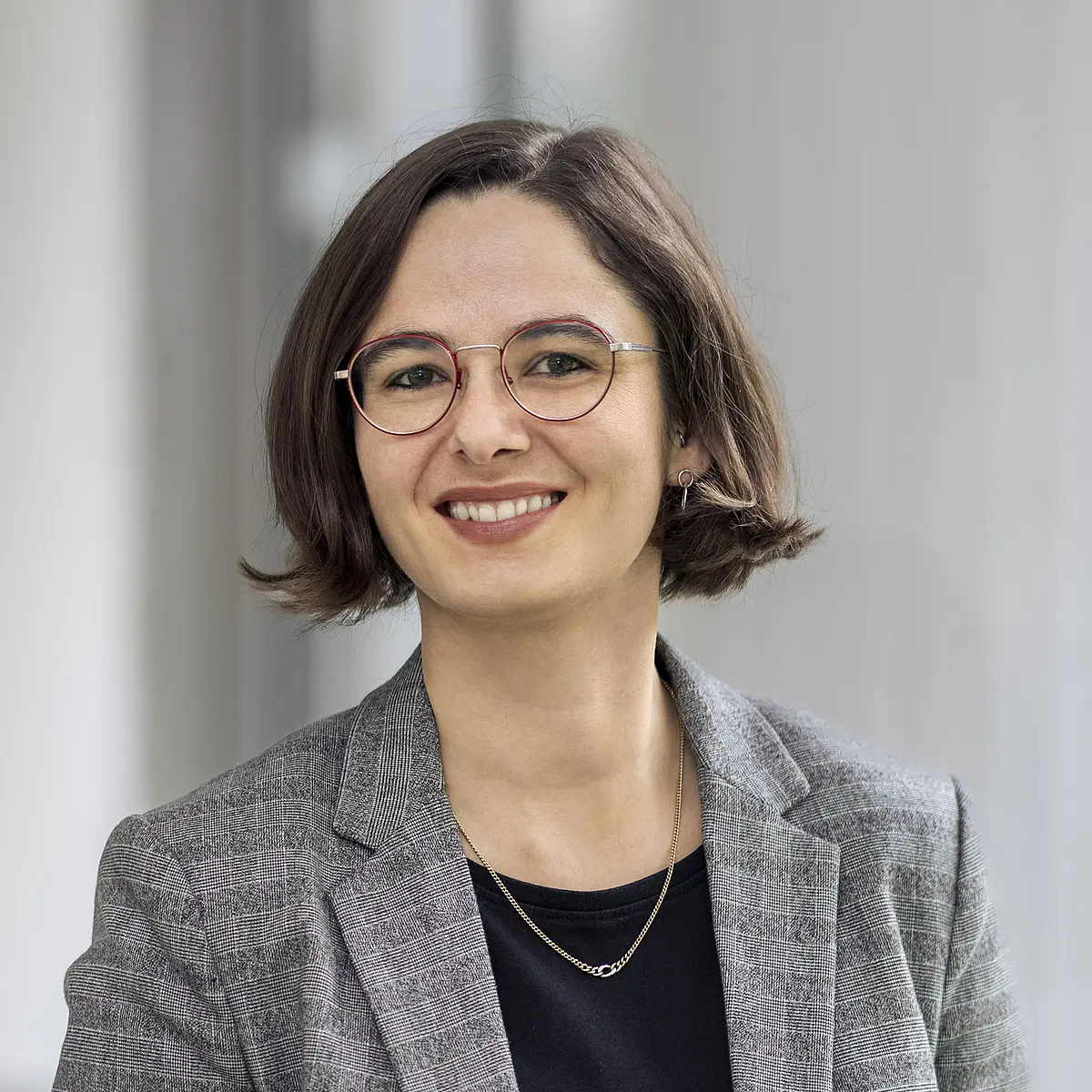 Professional portrait of Leonie Bossert, a white woman with a dark brown bob hairstyle wearing red, oval glasses, black and white glenckeck blazer over a black shirt with a simple silver chain necklace, smiling into the camera. 