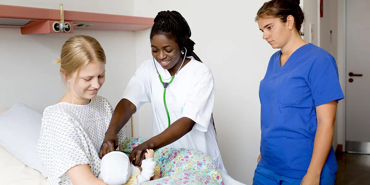 Eine junge Hebamme untersucht ein neugeborenes Kind mit einem Stethoskop. Das Kind liegt dabei in den Armen seiner Mutter in einem Krankenbett. 