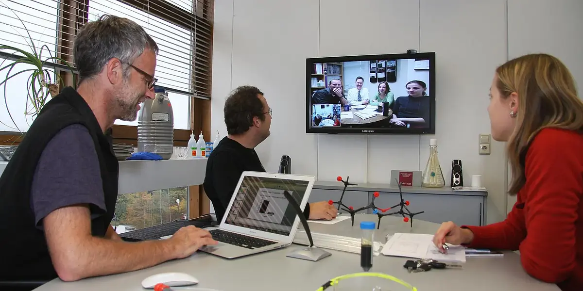 Three researchers at a table during a video conference, other participants are seen on a screen on the wallm Bildschirm an der Wand zu sehen