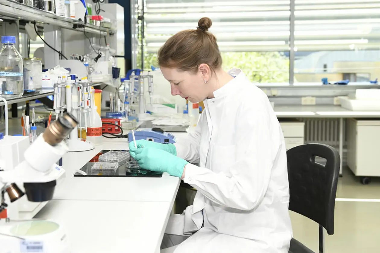 Dr. Deborah Kronenberg-Versteeg sits in a white coat and turquoise gloves in a laboratory containing various instruments. She is working in a concentrated manner.