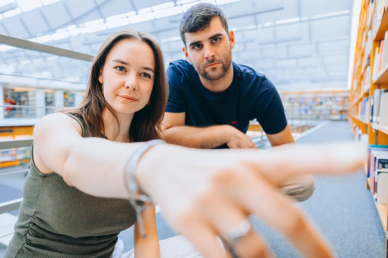 Eine Studentin zeigt auf ein Buch in der Bibliothek. Ein Student steht neben ihr und schaut ihrem Finger nach.