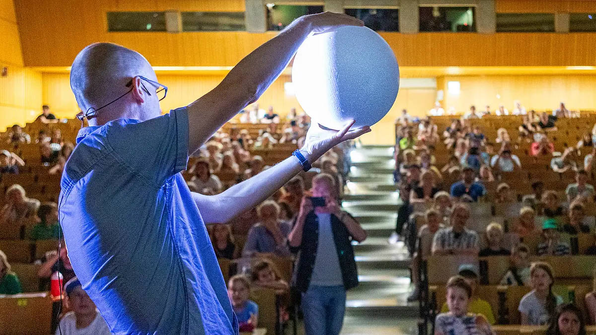 Ein Dozent hält eine hell beleuchtete Kugel in die Luft und präsentiert sie den Kindern im Hörsaal. Die Kugel wird angeleuchet, wie der Mond.