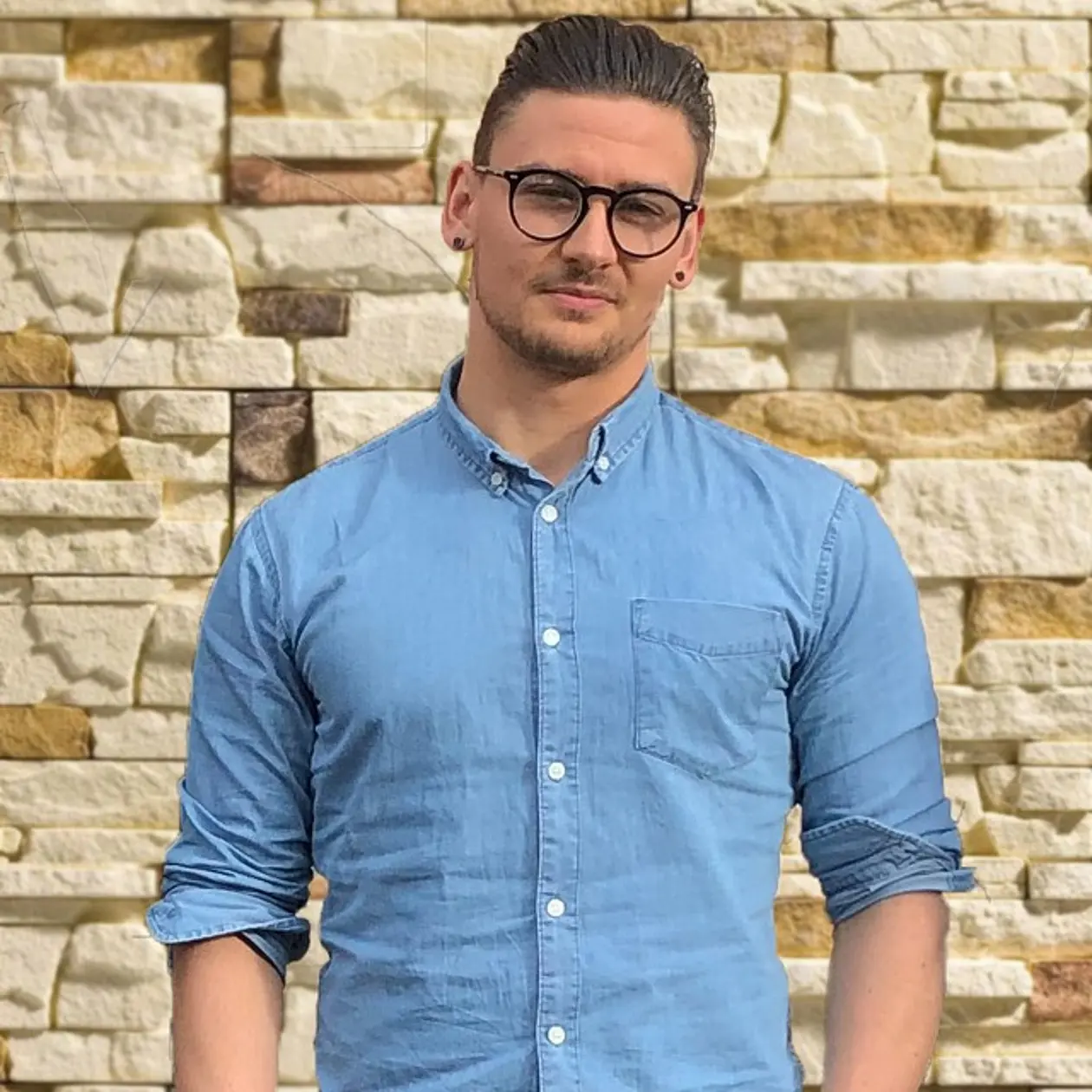 The image shows David Boysen, a white man with short dark hair and black framed glasses. He wears a short beard and a light blue button down shirt as well as black studded earrings. He is standing in front of a light brown stone brick wall with his head slightly bend to the left. 