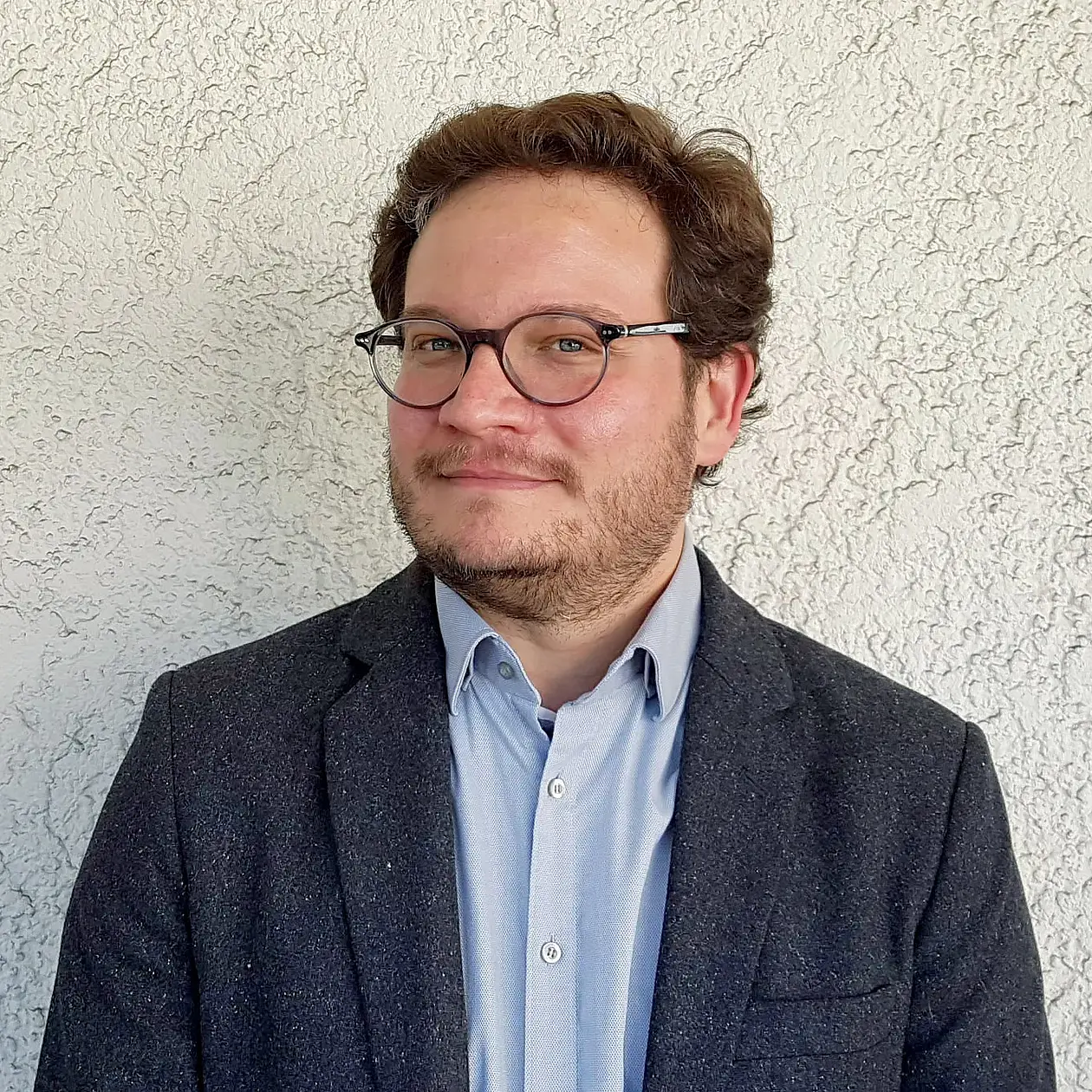 An image of Christopher Miller, a white amn with short wavy dark hair, with glasses and in a light blue button down shirt under a dark blue blazer, standing in front of a light grey wall. 