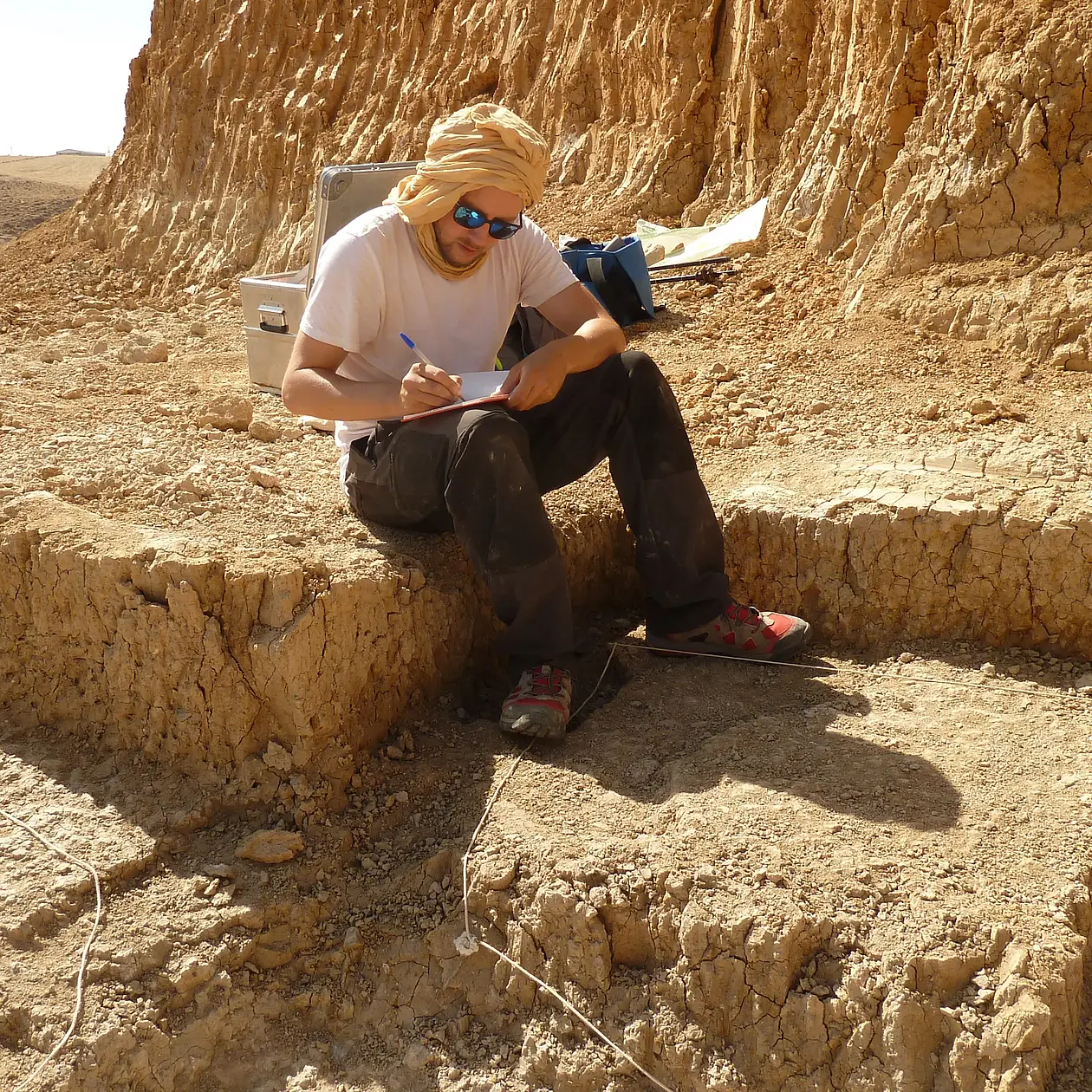 Mathieu Duval is a white man, in this photo sitting on the rim of a pit on an excavation site. He is wearing heavy dark cargo trousers, a white shirt and a beige shawl he has wrapped around his head and neck as well as dark sunglasses. He is writing in a journal. 