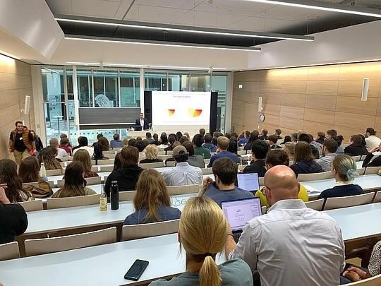 Gefüllter Hörsaal von hinten über besetzte Stuhlreihen bis hin zum Vortragenden fotografiert.