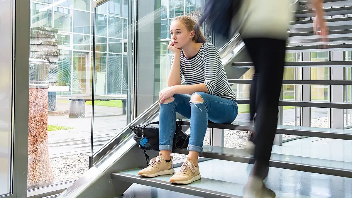 Studentin mit traurigem Blick sitzt auf einer Treppe, andere Personen gehen an ihr vorbei