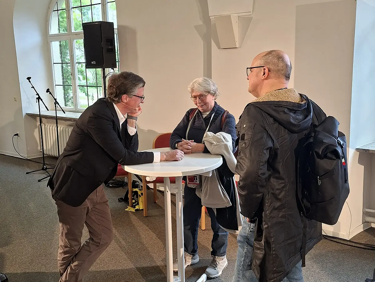 Auf diesem Bild sind drei Personen zu sehen, die sich um einen Stehtisch arrangiert haben. Professor Winter steht auf der linken Seite des Tisches. Ihn sieht man im Profil. Er schmunzelt leicht. In der Mitte des Bildes ist eine ältere Frau zu sehen. Sie ist klein, trägt eine Brille und hat den Kopf leicht geneigt. Auch sie schmunzelt leicht. Auf der rechten Seite des Bildes steht ein Mann mit Glatze. Sein Gesicht ist nicht zu sehen.
