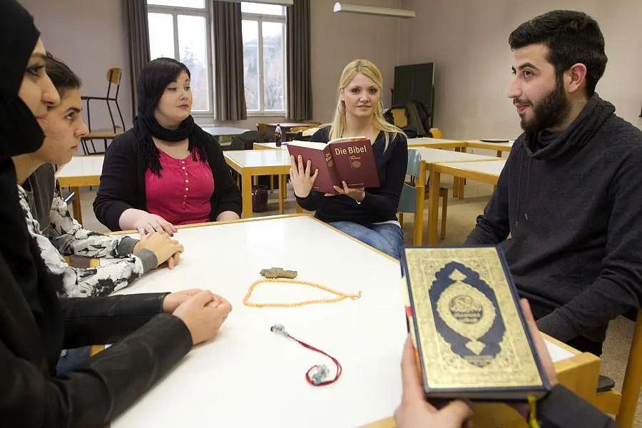 Sechs Studierende befinden sich in einem Seminarraum und sitzen um einen Tisch. Eine junge Frau hält die Bibel in der Hand, eine andere Person, von der nur die Hände zu sehen sind, hält den Koran.