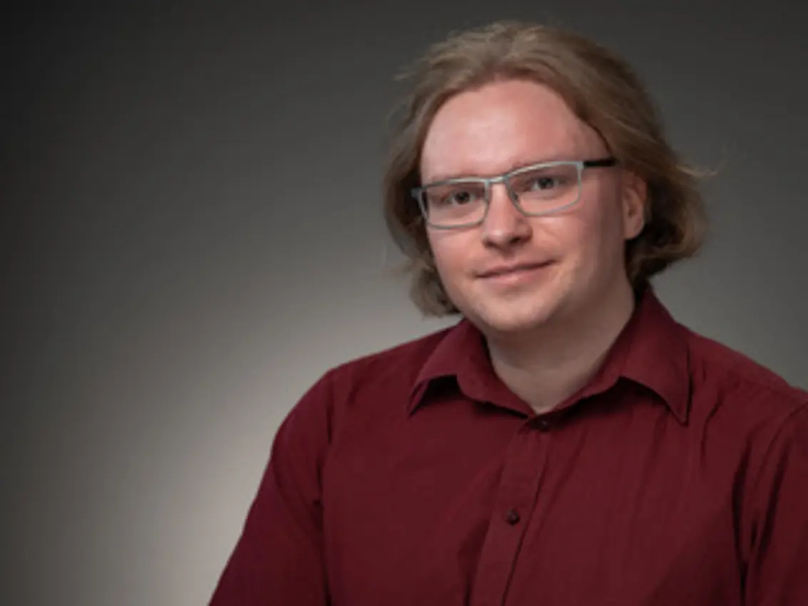 A professional photoshoot of Stefan Greiner, a white man with ginger medium long hair and rectangular glasses. He wears a dark read button down shirt. 