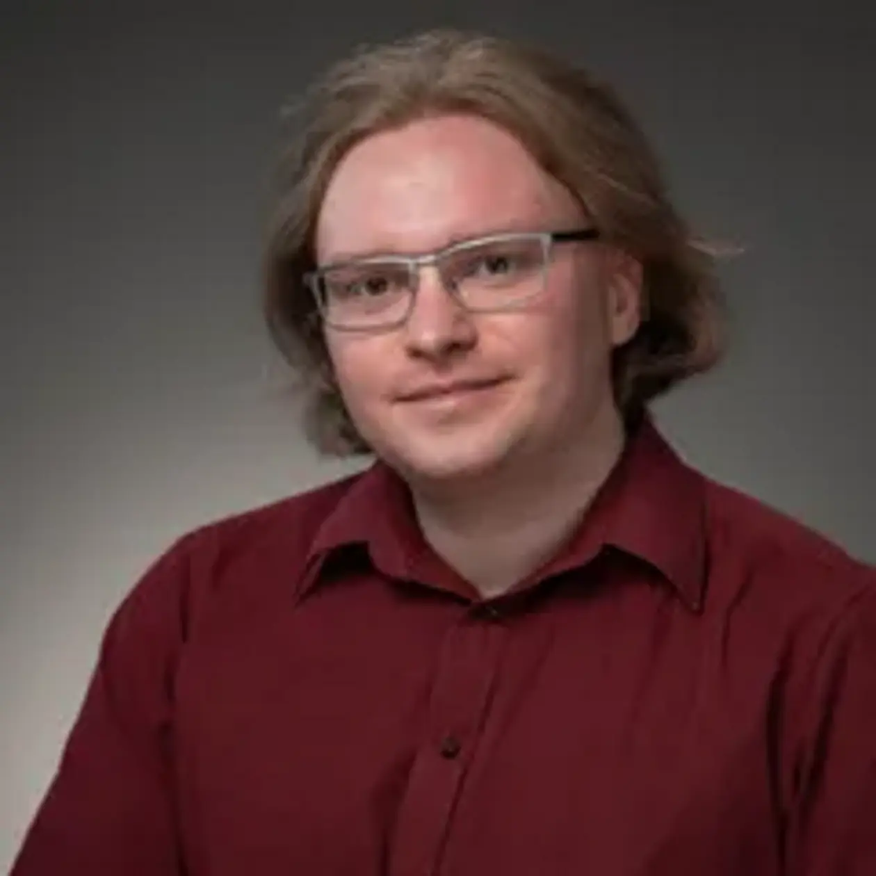 A professional photoshoot of Stefan Greiner, a white man with ginger medium long hair and rectangular glasses. He wears a dark read button down shirt. 