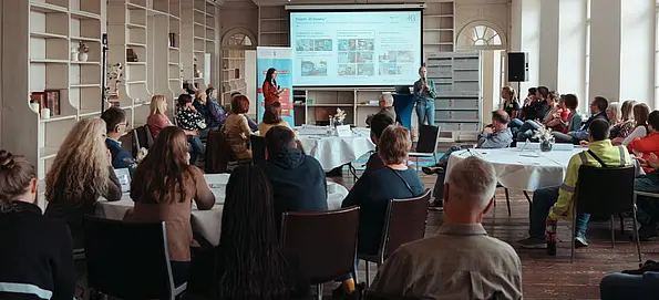 Etwa 30 Personen sitzen in Gruppen an großen Rundtischen mit dem Rücken zur Kamera. Im Bildhintergrund stehen 2 Personen vor einer Leinwand, auf der eine Powerpointpräsentation erkennbar ist, und halten einen Vortrag für die Anwesenden.