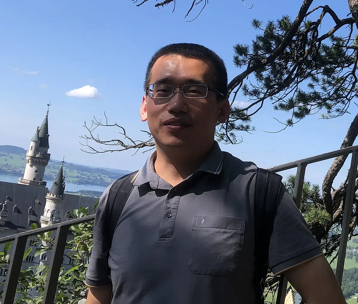 Aufnahme von Yingxin Li, der vor einem Metallgeländer steht und in die Kamera lächelt. Im Hintergrund ist ein weißes Schloss mit Türmen in einerr Landschaft aus Felder, Seen, und Siedlungen zu sehen.