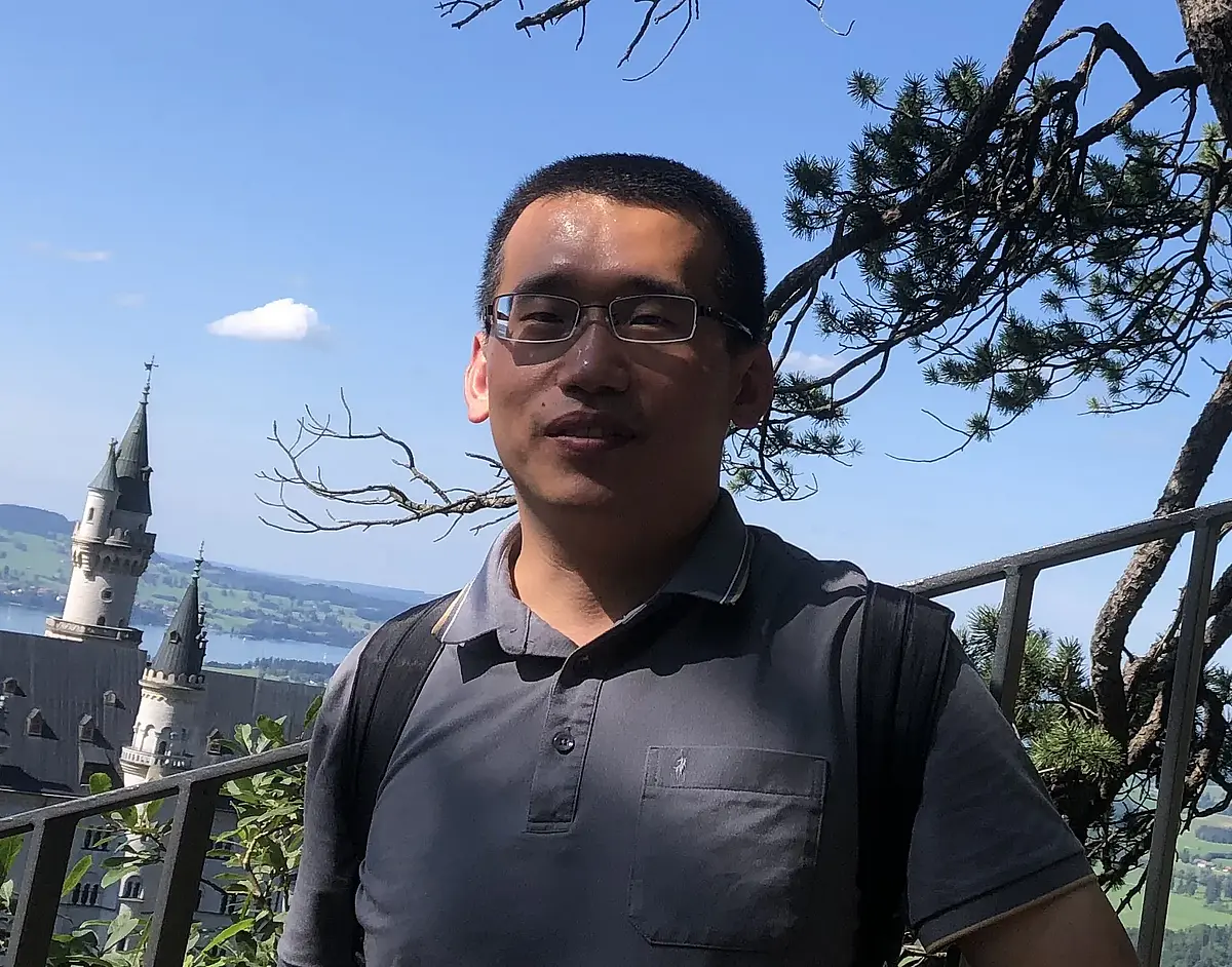 Photography of Yingxin Li standing in front of a metal railing and smiling at the camera. In the background is a white castle with towers in a landscape of fields, lakes and settlements.
