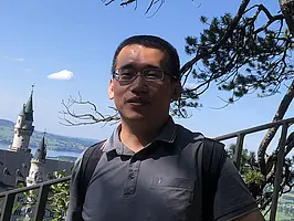 Aufnahme von Yingxin Li, der vor einem Metallgeländer steht und in die Kamera lächelt. Im Hintergrund ist ein weißes Schloss mit Türmen in einerr Landschaft aus Felder, Seen, und Siedlungen zu sehen.
