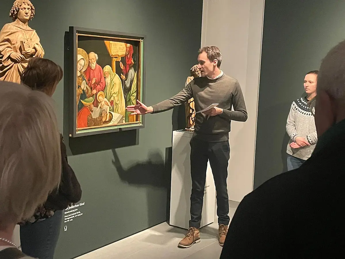 Auf dem Bild ist ein junger Mann im Rottenburger Diözesanmuseum zu sehen, der vor einem Gemälde steht, welches Jesu Beschneidung zeigt. Das Gemälde hängt an einer dunkelgrünen Wand. Um den jungen Mann stehen Zuhörende unterschiedlichen Alters.