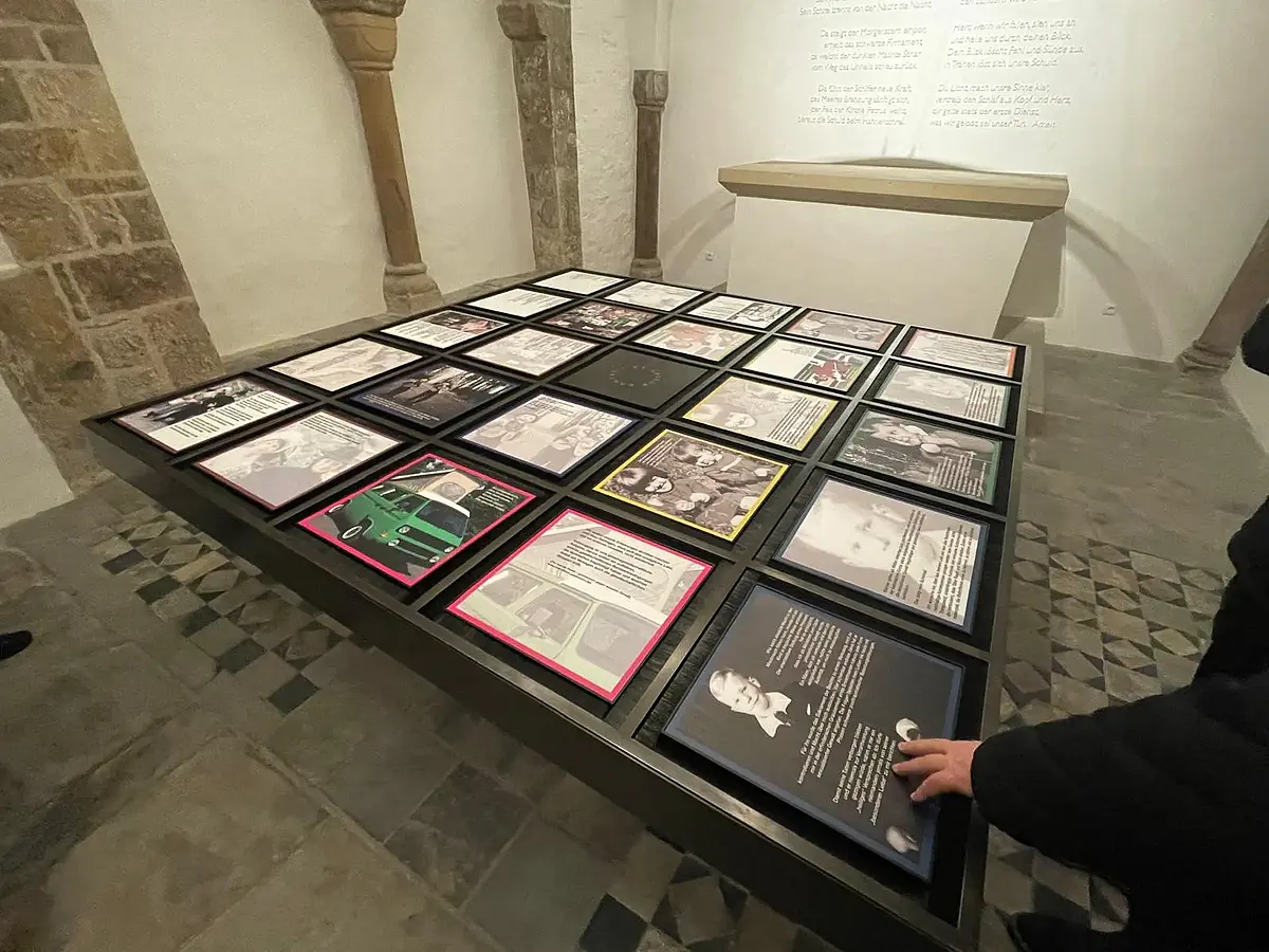 zu sehen ist ein quadratischer schwarzer Tisch in der Mitte eines Krypta-ähnlichen Raums. Auf dem Tisch befinden sich quadratische Bildrahmen. Sie zeigen z.T. Porträtfotos aber auch kurze Zitate.