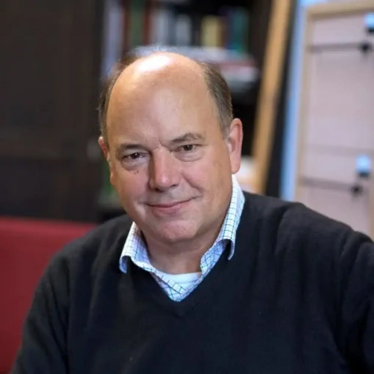 Photo of Professor Nicholas Conard. He is a white man in his sixties with a mostly bald head, sitting facing the camera, wearing a dark blue sweater and a white collared shirt underneath, facing the camera smiling with hands folded in front of his torso. 