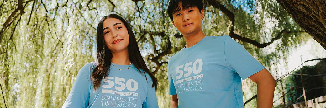A man and a woman wearing light blue 550th-anniversary T-shirts. There are trees in the background.