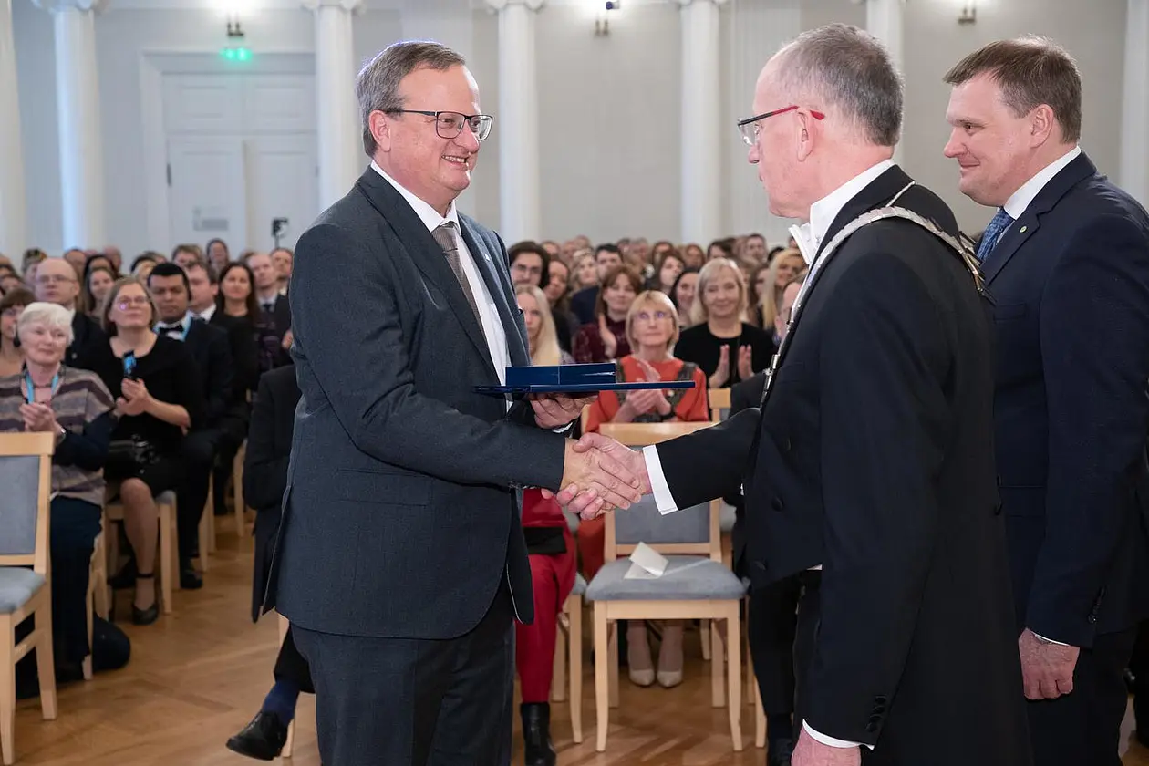 Harald Baayen and Toomas Asser shake hands in front of a large audience, Anti Selart is standing next to them