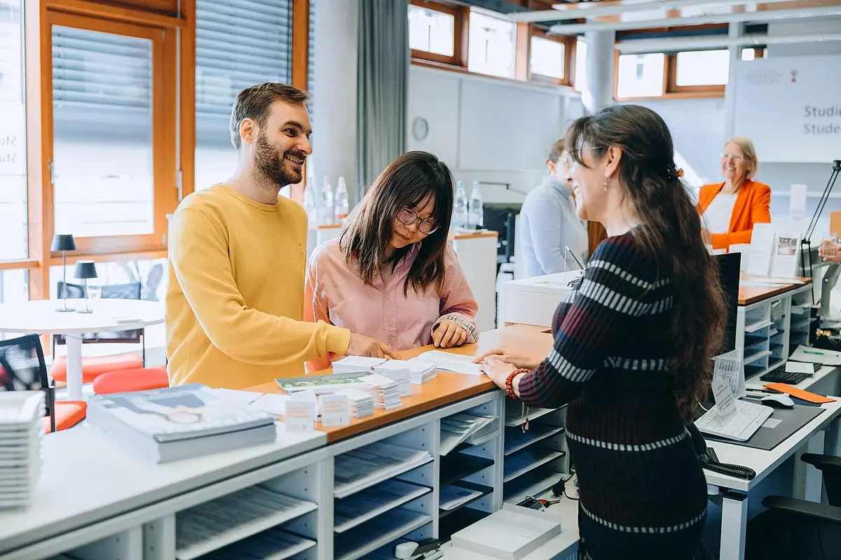 Zwei Studierenden stehen an einer Theke im Gespräch mit einer Mitarbeiterin hinter der Theke