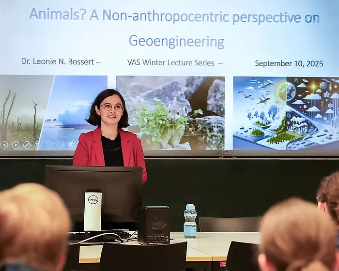 Leonie Bossert, a white woman with a dark brown bob hairstyle wearing oval glasses and a cherry red blazer over a black shirt, is presenting a talk. She stands behind a desk with a computer screen and a bottle of water in front of her, there are heads of the audience in the foreground. In the background, a large digital screen shot the title slide of her presentation, stating: Animals? A Non-anthropcentric perspective on Geoengineering. Dr. Leonie N. Bossert - VAS Winter Lecture Series - September 10, 2025. 