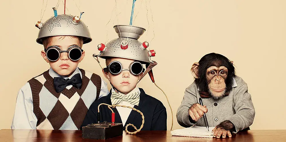 Two children with dark glasses wear devices made from kitchen sieves with propellers on their heads, which are connected to a black box via cables. Sitting next to them is a clothed chimpanzee who appears to be taking notes on a pad.