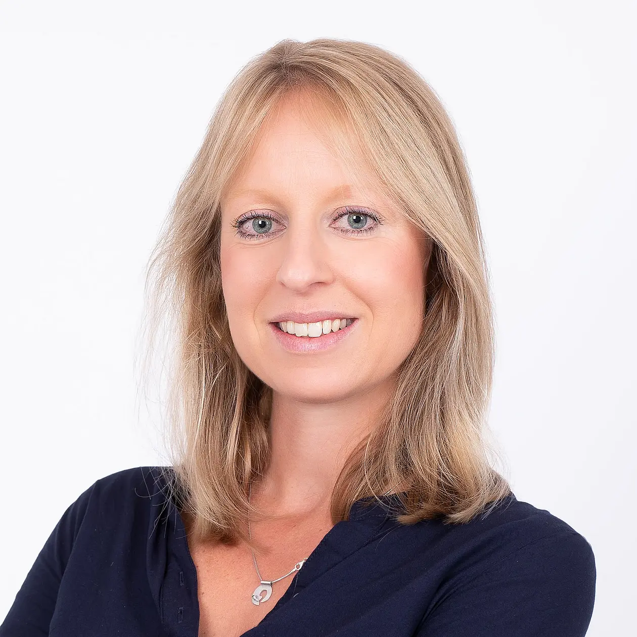 A portrait of Marlen Fröhlich, a white woman with shoulderlenght blond hair, smiling into the camera, wearing a dark blue blouse and a silver necklace with a pendant. 