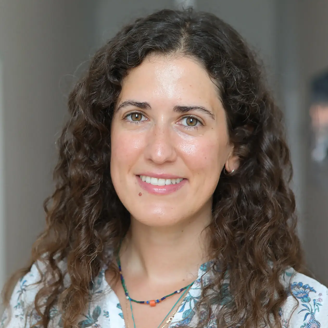 A portrait photo of Maria Spyrou, a white woman with long dark brown curly hair smiling into the camera. She is wearing a button down shirt with flowers and a chunky golden necklace.