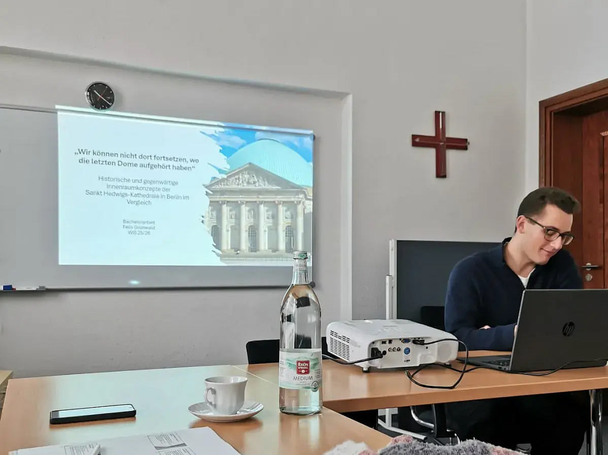 Zu sehen ist ein junger Student mit dunkelblonen Haaren und Brille, der sich am rechten Bildrand befindet. Den Mittelpunkt des Bilds bildet eine Tafel auf die eine Präsentation projiziert wird.