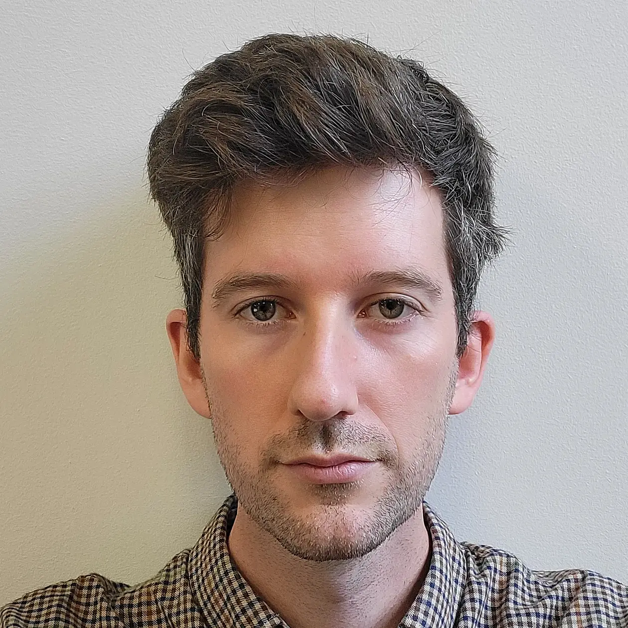 A portrait photo of Jaap Sears, a white man with short dark brown hair with grey, facing the camera in a serious manner, wearing a button down shirt with a brown and beige checkerboard pattern