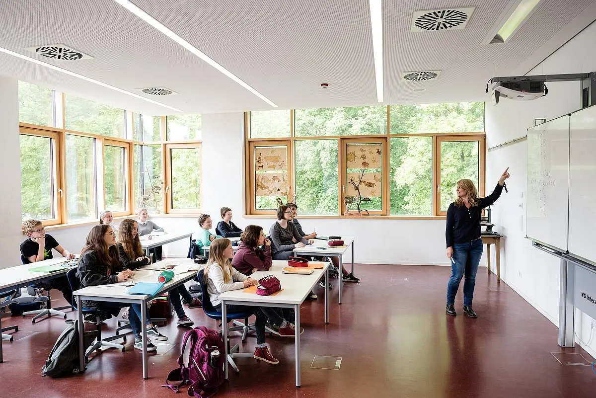 Schülerinnen und Schüler mit Lehrerin im Klassenzimmer. Die Lehrerin zeigt etwas an der Tafel.
