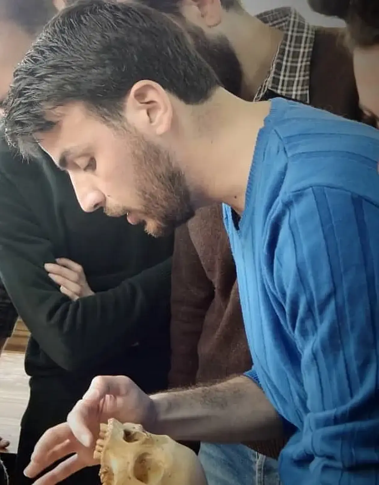 Photo showing Majid Aljaber Abo Fakher, a man with short brown hair and a short brown beard wearing a blue sweater holding a human skull and inspecting the teeth of the individual. 