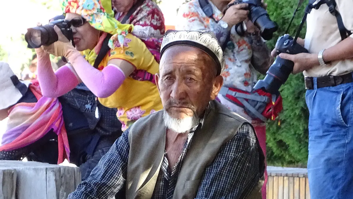 Ein trauriger älterer Mann mit uighurischer Kopfbedeckung und fotografierenden Han-Chinesen im Hintergrund