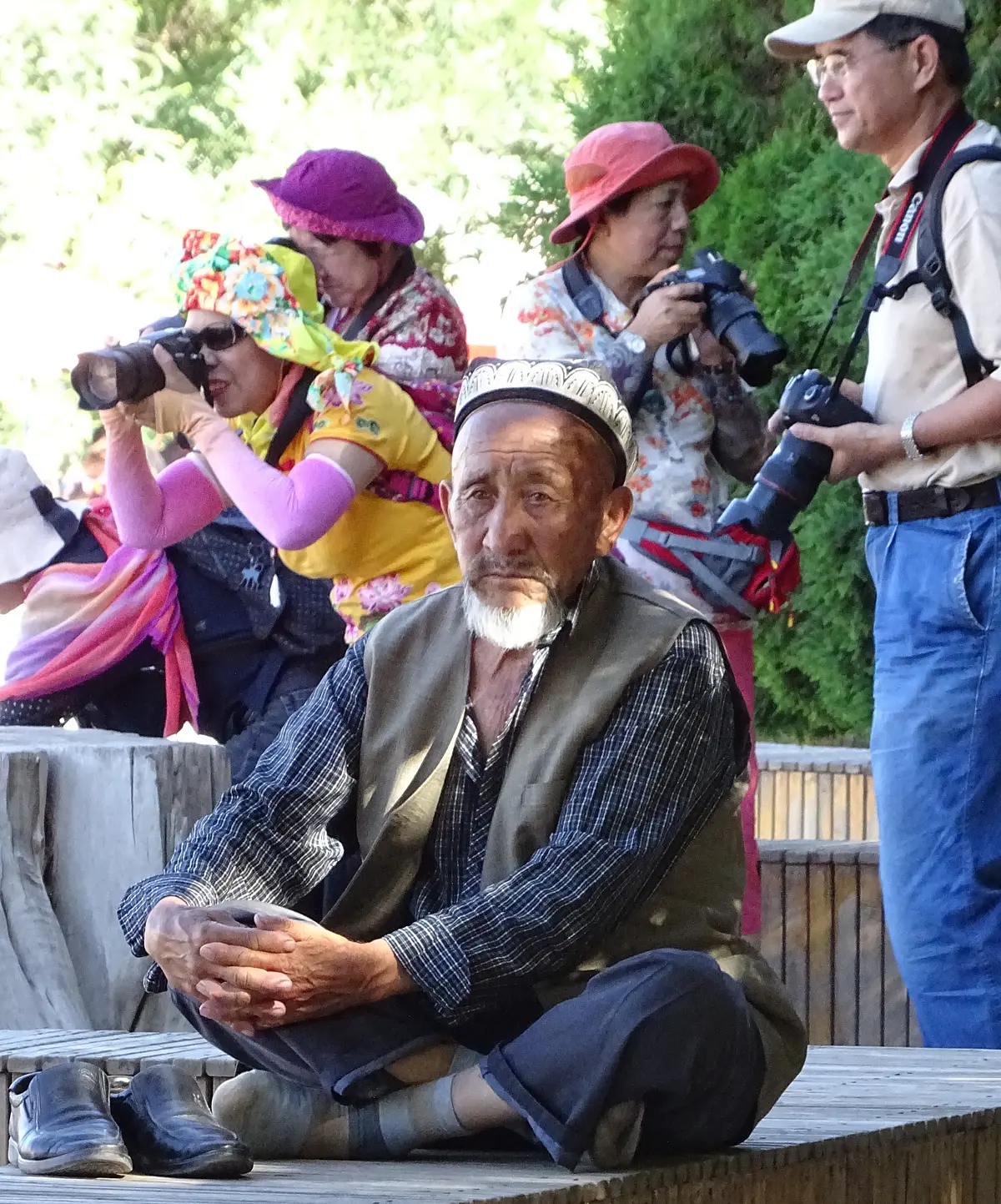 A sad elderly man with Uyghur headgear and photographing Han Chinese in the background