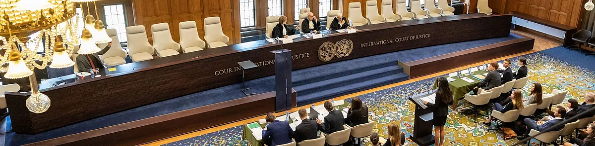 Aufnahme einer Verhandlung im Friedenspalast in Den Haag. Zu sehen sind drei Richter, die an der Richterbank Platz genommen haben und eine Teilnehmerin, die - an einem Pult stehend - ihr Plädoyer hält.  