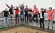 The research group of Vision and Cognition stands outside on a bannister in front of mountains. Some members have their hands up in the air.