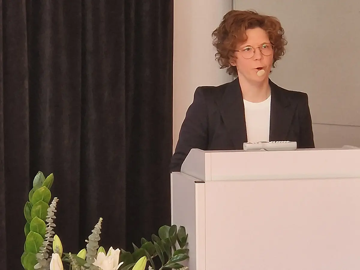 in der rechten Bildhälfte ist Lisa Kühn zu sehen, die ein schwarzes Jackette und eine weiße Bluse trägt. Sie steht hinter einem weißen Ambo. In der linken Bildhälfte ist ein sehr großer Blumenstrauß zu sehen. Der Hintergrund wird überwiegend von einem schwarzen Vorhang eingenommen.