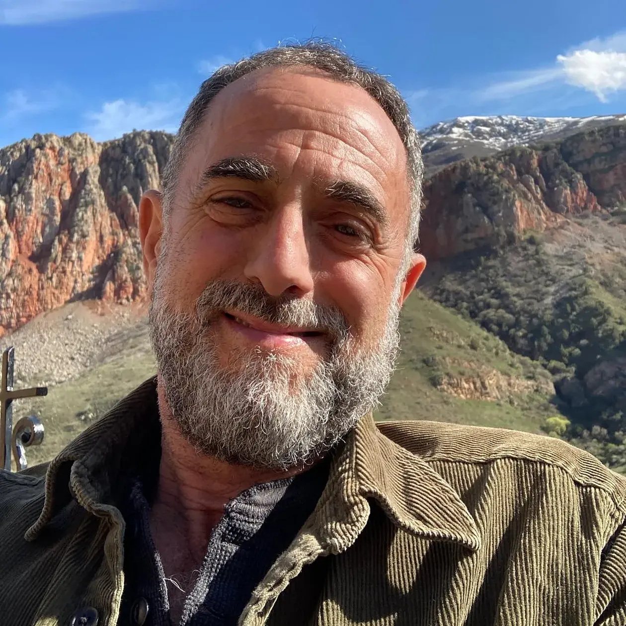 A selfie of Andrew Kandel, a white man with very short black and grey hair and a short black and grey beard, wearing a olive corduary jacket smiling widely in front of a reddish mountain range under blue sky.
