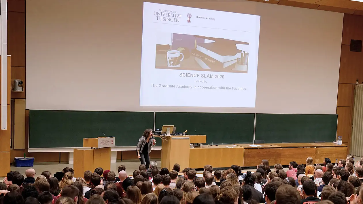 Picture from the Science Slam 2020. A full lecture hall can be seen, the moderator gives a speech and a presentation with the graphic of the doctoral days days can be seen.